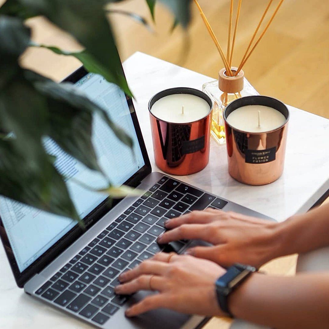 Zwei Duftkerzen neben einem Laptop und einem Diffusor. Kerzen mit Aufschrift: Atelier Rebul, Flower Fusion. Hände tippen auf der Tastatur.