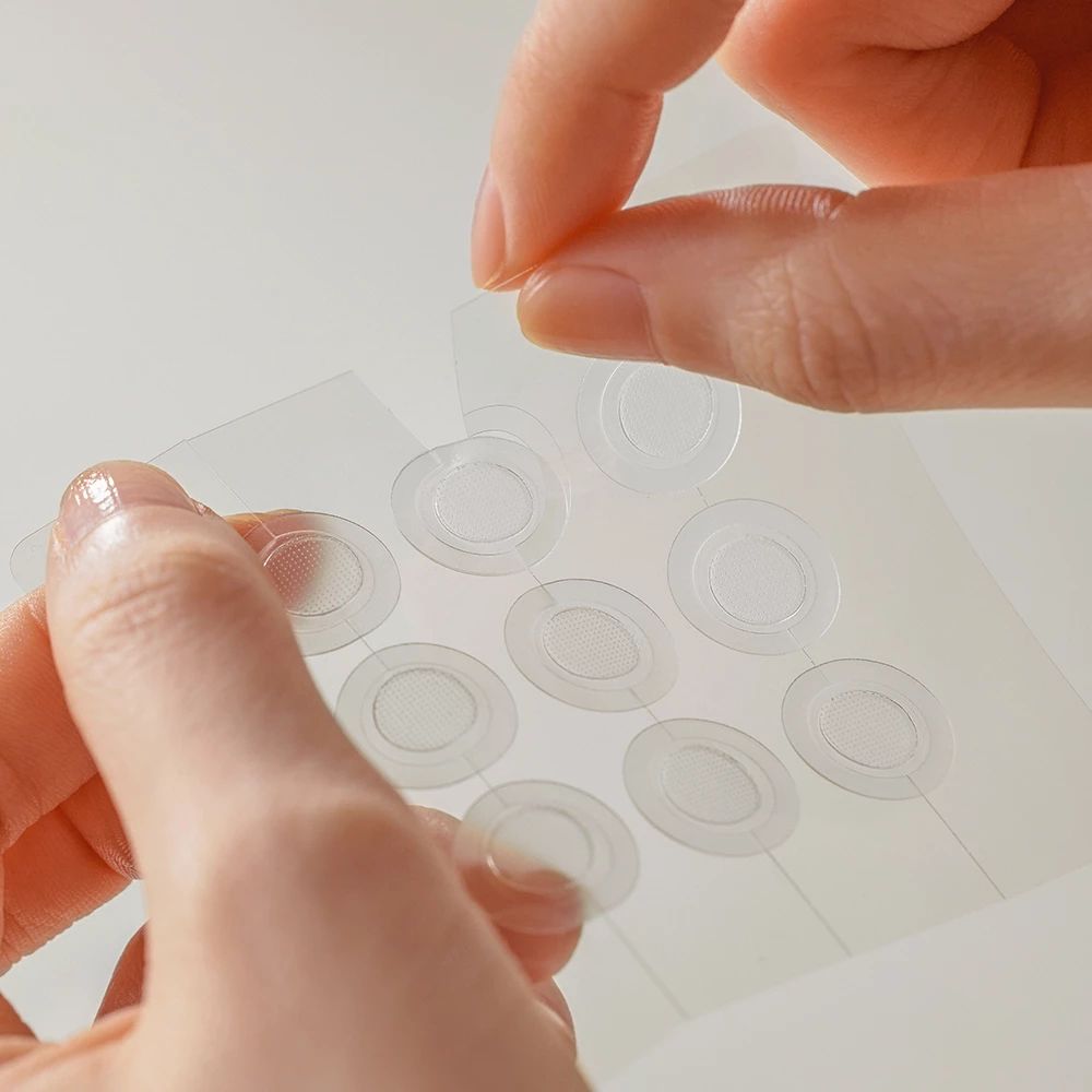 Hände entfernen transparente, runde Patches von einer transparenten Folie. 10 Patches sichtbar.