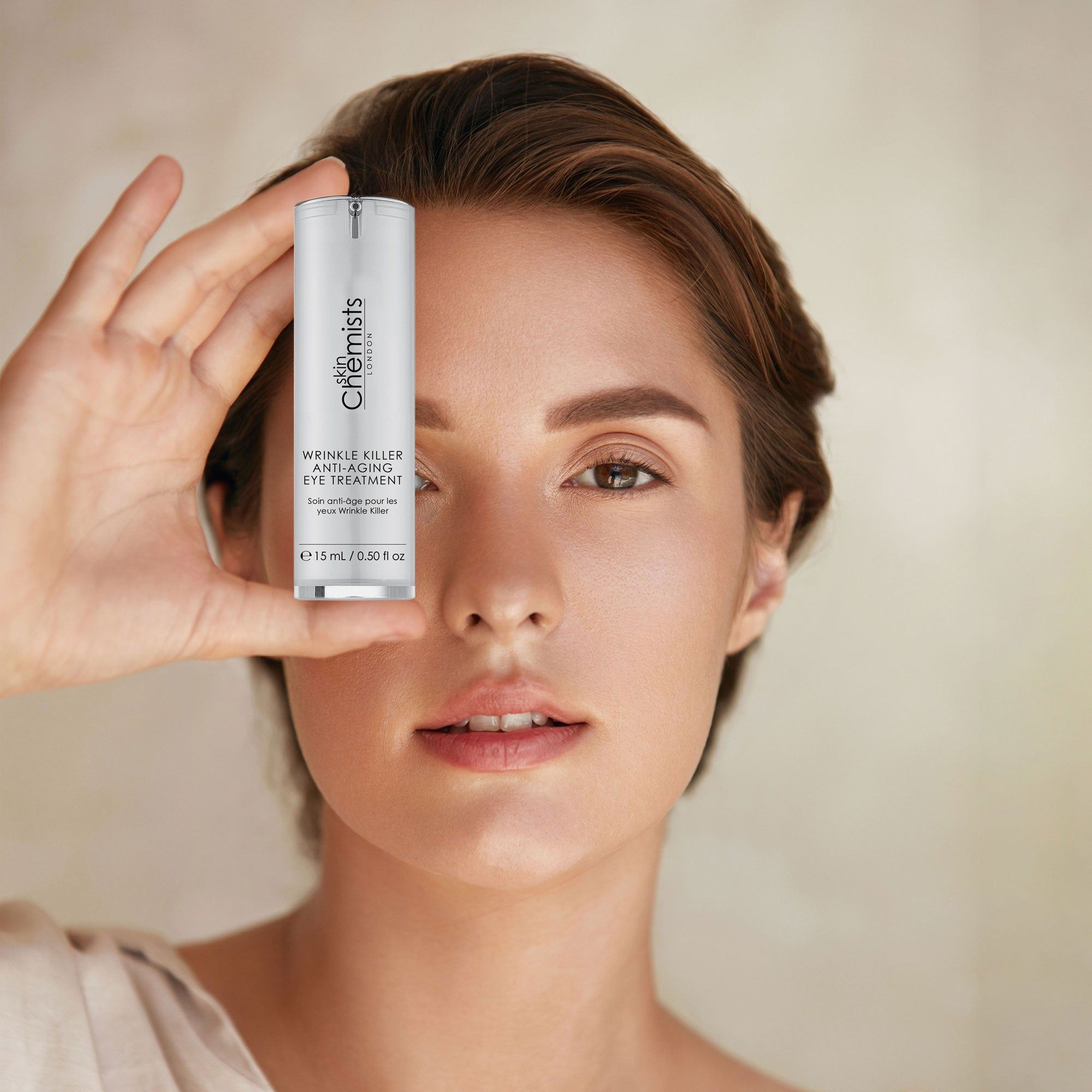 Frau hält zylindrische Augenbehandlung vor dem Auge. Weißer Behälter mit silbernem Deckel. Text: Wrinkle Killer Anti-Aging Eye Treatment, 15 ml / 0.50 fl oz.