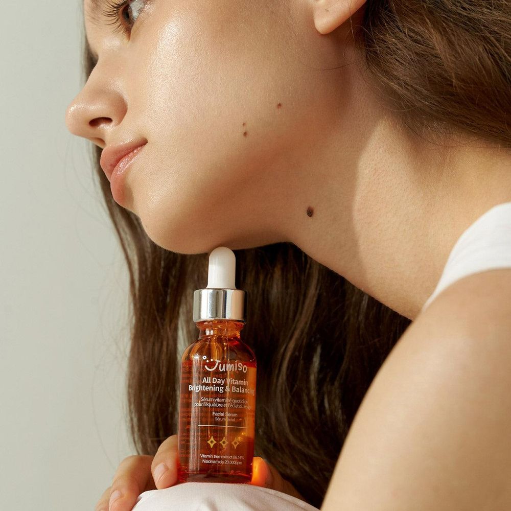 Frau hält eine Flasche Serum. Aufschrift: JUMISO All Day Vitamin Brightening & Balancing. Gesicht im Profil.