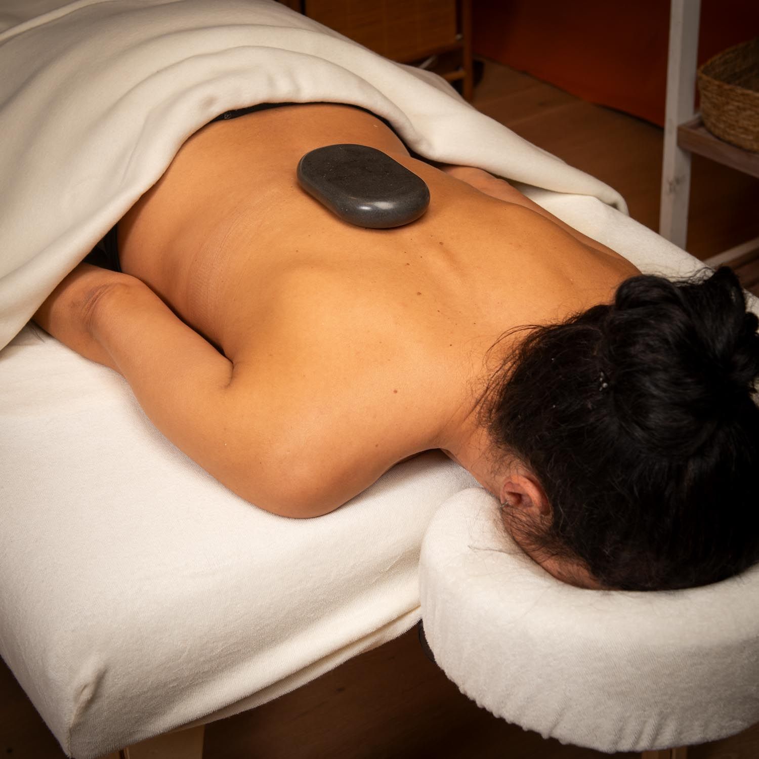 Großer, ovaler Stein auf dem Rücken einer Person. Person liegt auf einer Liege, bedeckt mit einem weißen Tuch.