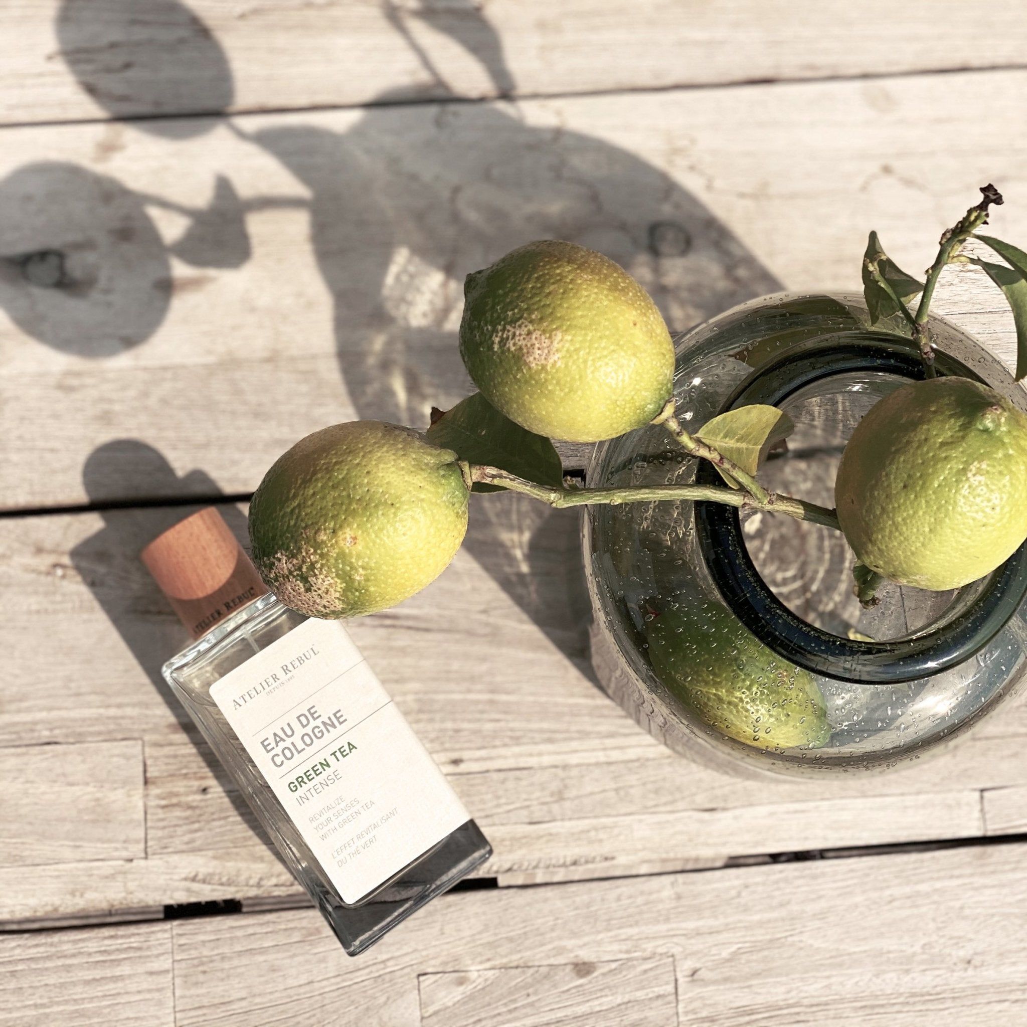 Glasflakon mit Holzverschluss. Aufschrift: Atelier Rebul, Eau de Cologne Green Tea Intense. Neben Zitronen und Zweigen in Glasvase.