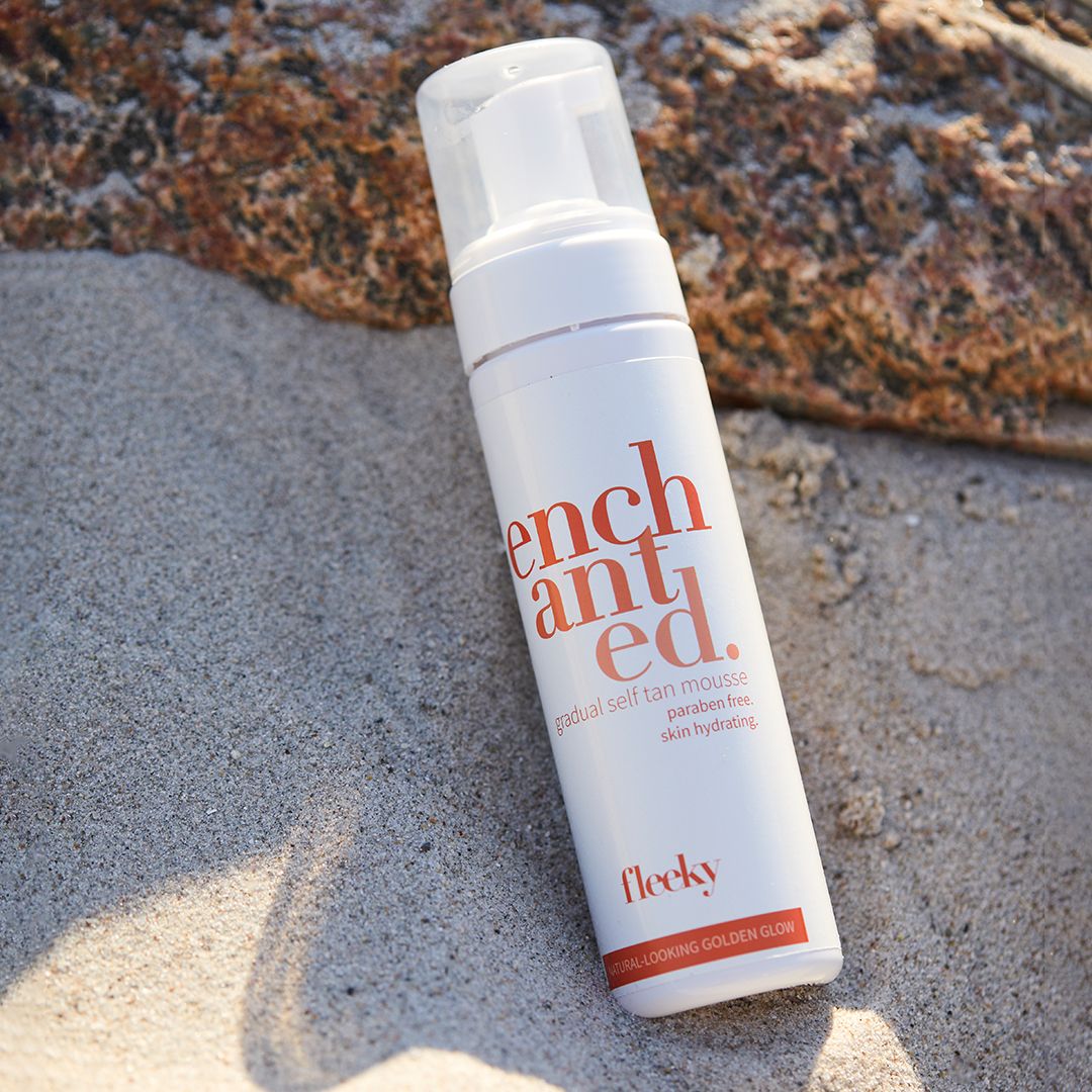 Weißes Mousse-Behältnis auf Sand und Stein. Aufschrift: enchanted. Gradual Self Tan Mousse. Marke: fleeky.