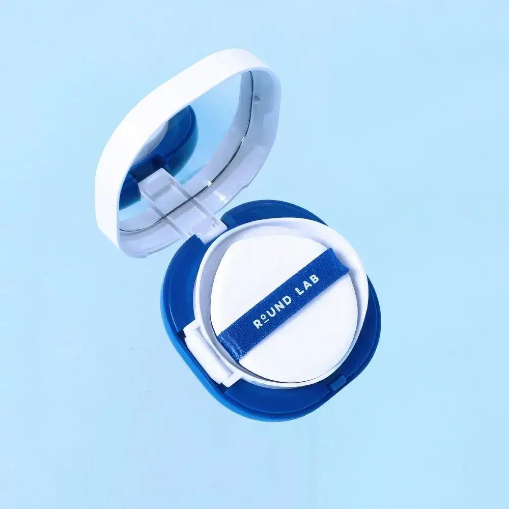 Blau-weiße Kosmetikdose mit Spiegel und Schwamm. Schwamm mit blauem Band und Schriftzug ROUND LAB. Geöffnet.