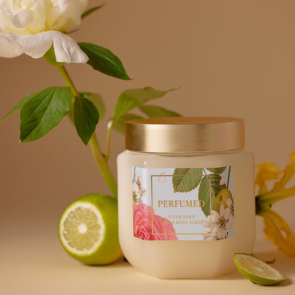 Glasbehälter mit goldfarbenem Deckel und Etikett, neben einer Limette und einer Blume. Aufschrift: PERFUMED FLEUR SOAP SUGAR BODY SCRUB.
