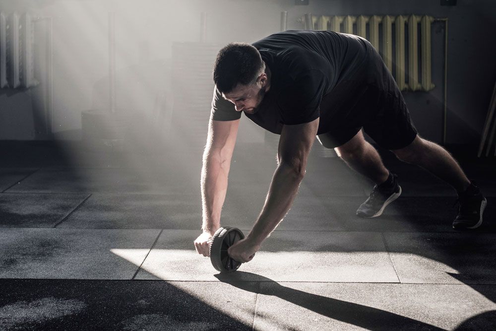 Mann trainiert mit Fitnessroller. Schwarzes Rad, Griffe. In Sporthalle. Lichtstrahlen. Auf dem Boden liegend.