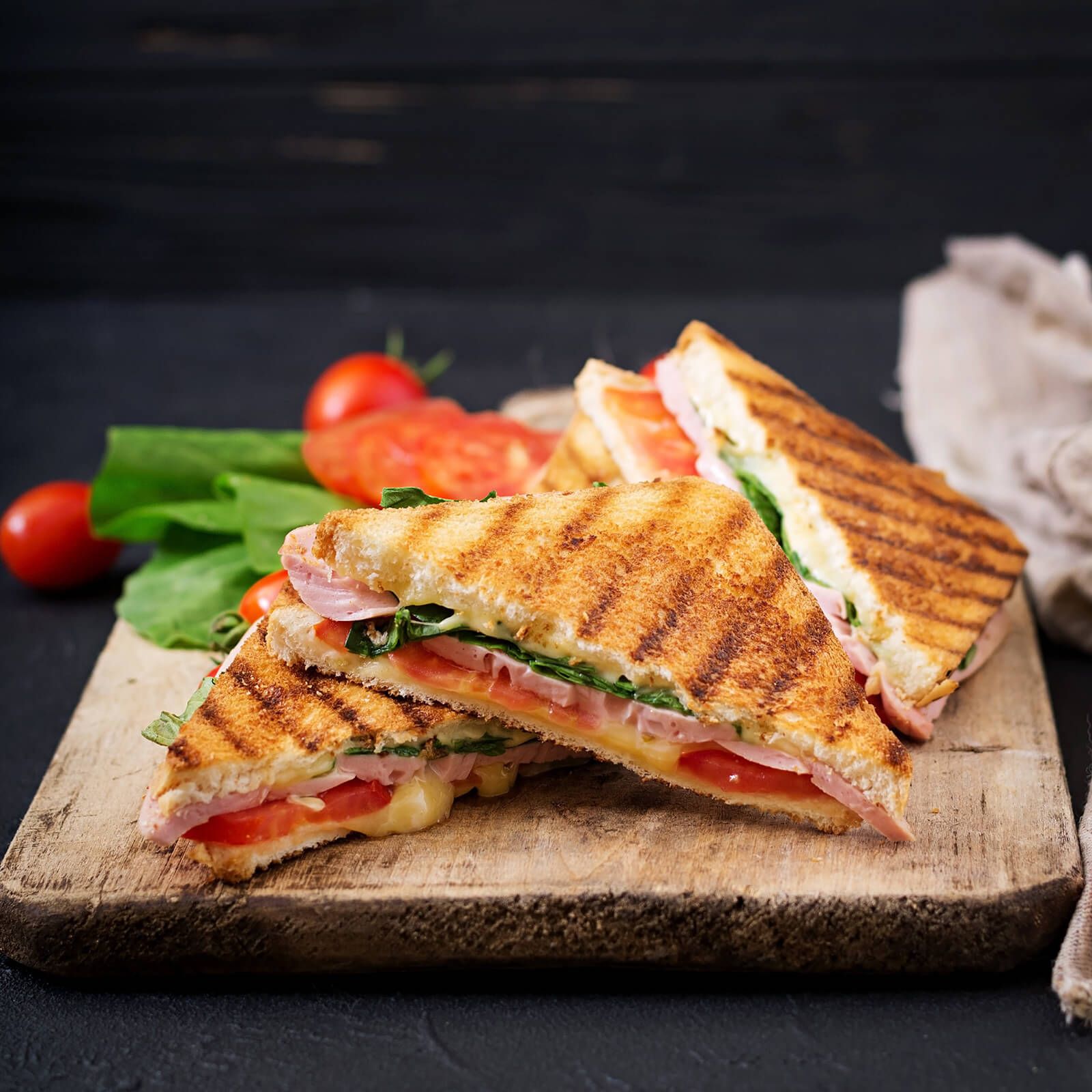 Gegrillte Sandwiches mit Käse, Schinken, Tomaten und Salat auf einem Holzbrett.