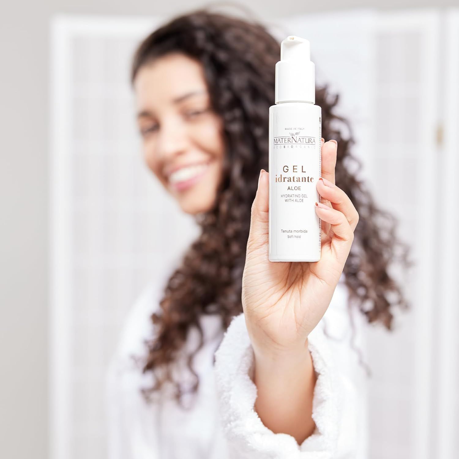 Frau mit Locken hält eine weiße Flasche mit Pumpspender. MATERNATURA, GEL idratante ALOE.