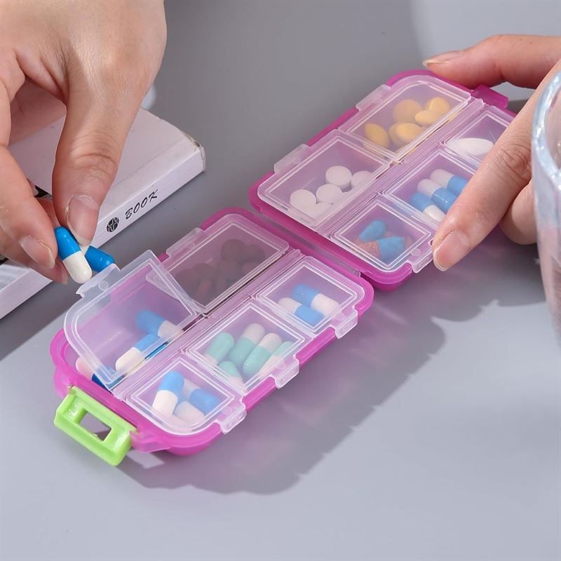 Pink Tablettenbox, geöffnet, mit sieben Fächern. Hände legen Pillen hinein. Grüner Griff.