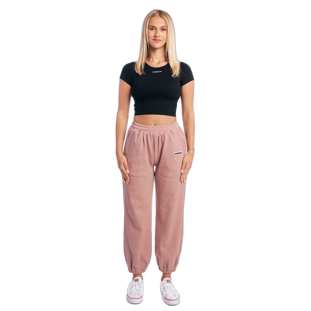 Frau in voller Länge trägt schwarzes Crop-Top und rosafarbene Jogginghose. Weißer Hintergrund.