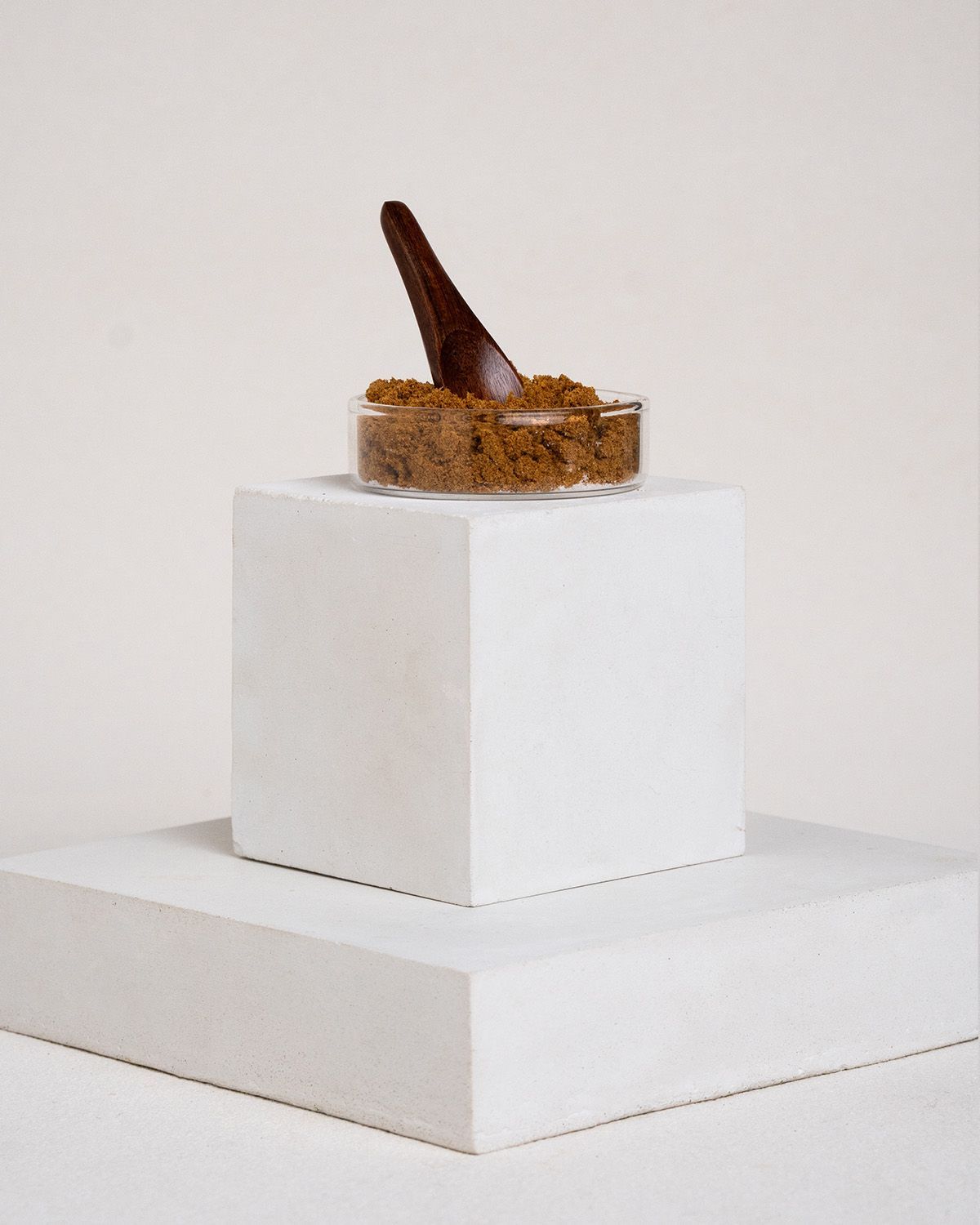Schale mit braunem Pulver, Holzlöffel. Auf weißem Podest.