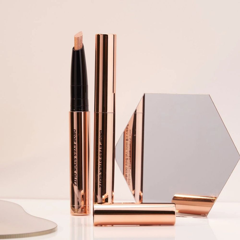 Mehrere roségoldfarbene Lidschattenstifte und ein Spiegel. Produktname: DROP EYES STICK GLITZ. Text auf den Stiften.