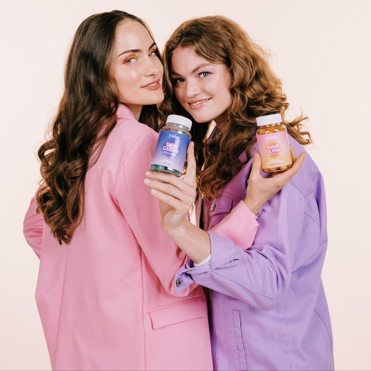Zwei Frauen halten jeweils eine Flasche Hair & Skin Vitamin Gummies Duo. Eine Frau hält eine blaue, die andere eine orangefarbene Flasche.