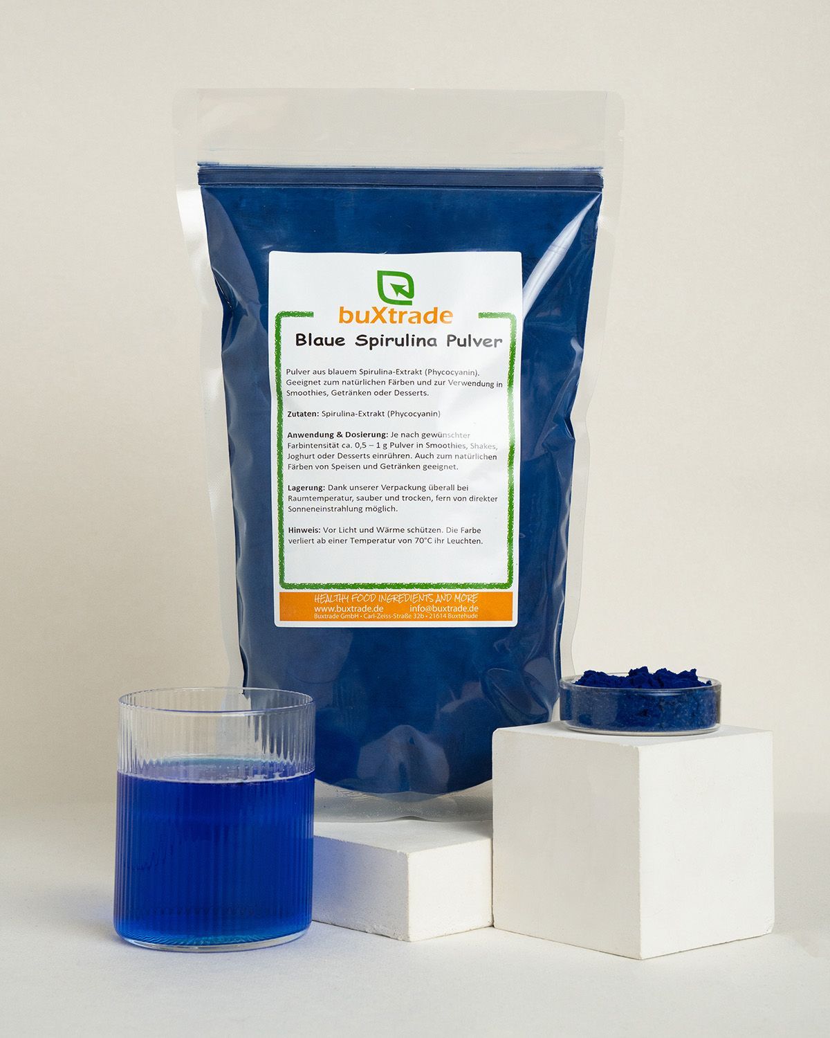 Buxtrade Blaue Spirulina Pulver in einem Standbeutel mit Etikett. Daneben ein Glas mit blauem Getränk und eine Schale mit Pulver.