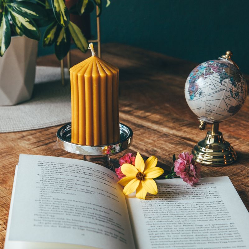 Gelbe Wachskerze auf silbernem Kerzenhalter. Auf einem Tisch mit Buch, Globus und Blumen dekoriert.