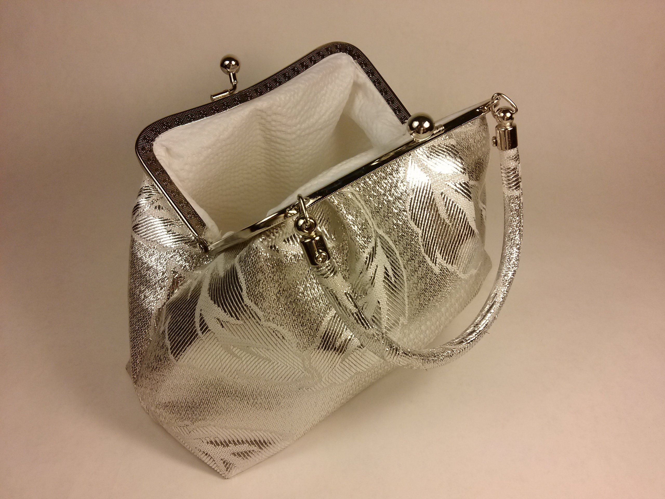 Silberfarbene Handtasche, geöffnet. Innenfutter weiß. Griff und Verschluss sichtbar.