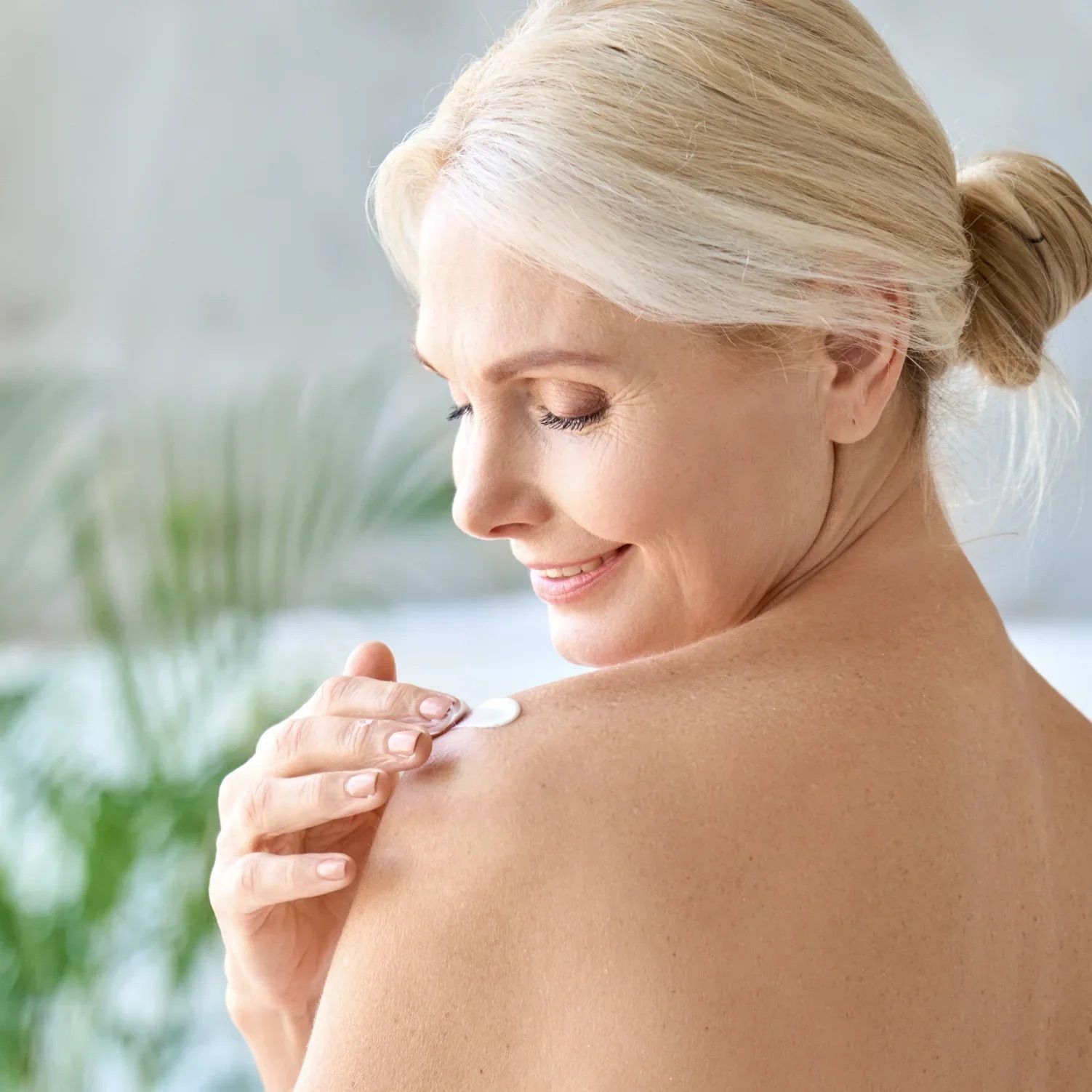 Frau trägt Körperlotion auf die Schulter auf. Haut ist sichtbar. Helles Badezimmer im Hintergrund.