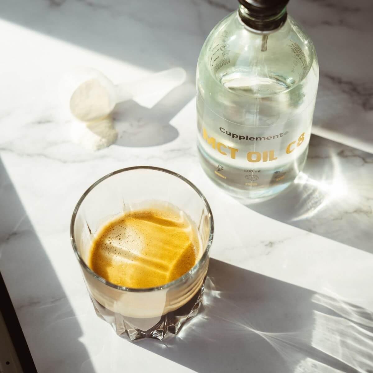 Glas mit Kaffee und MCT-Öl. Daneben eine Flasche MCT OIL C8. Aufschrift: ORGANIC. Auf hellem Untergrund.