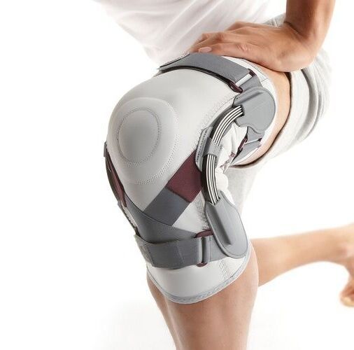 Person mit Kniebandage. Weiß-grau mit grauen Scharnieren und Riemen. Das Produkt ist am Knie angebracht.