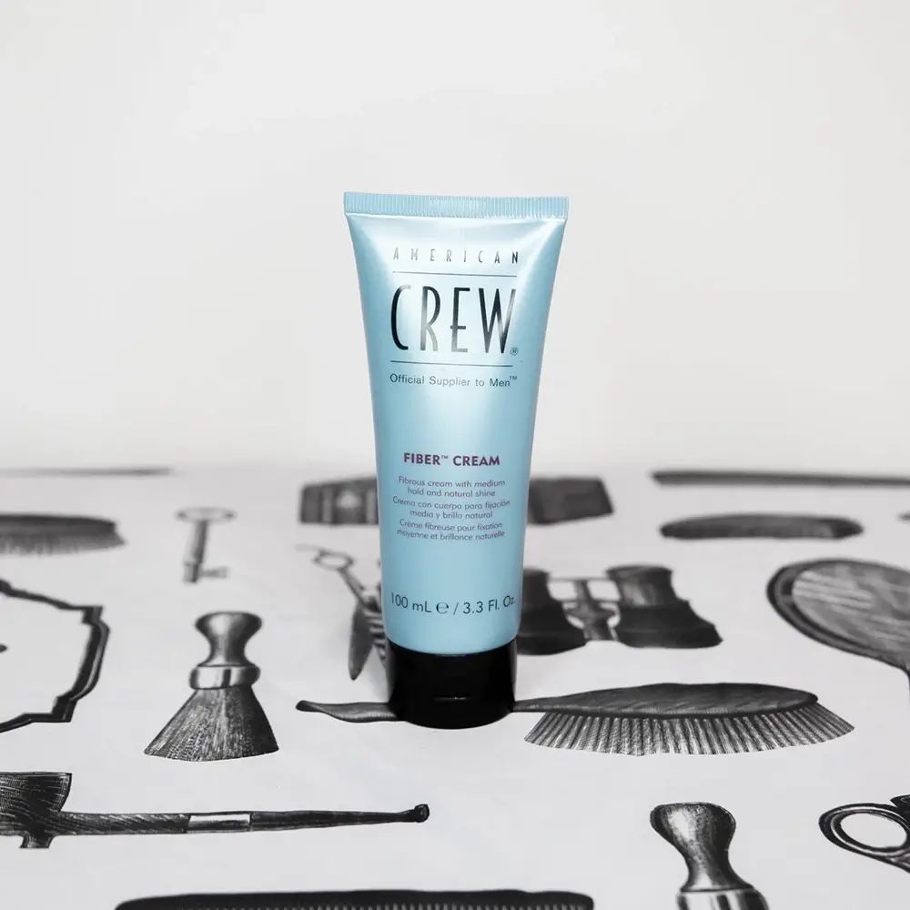 Hellblau Tube mit schwarzem Deckel. Aufschrift: American Crew, Fiber Cream. Auf Tisch mit Friseur-Motiven.