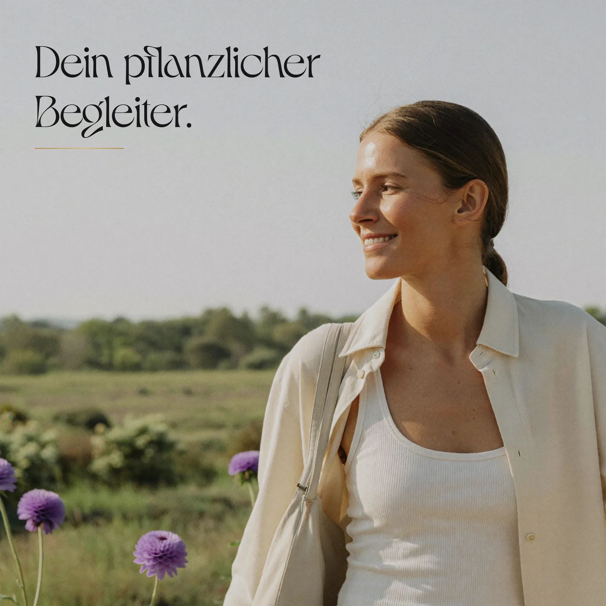 Frau in einem Feld mit lila Blumen, trägt ein weißes Tanktop und eine beigefarbene Bluse.