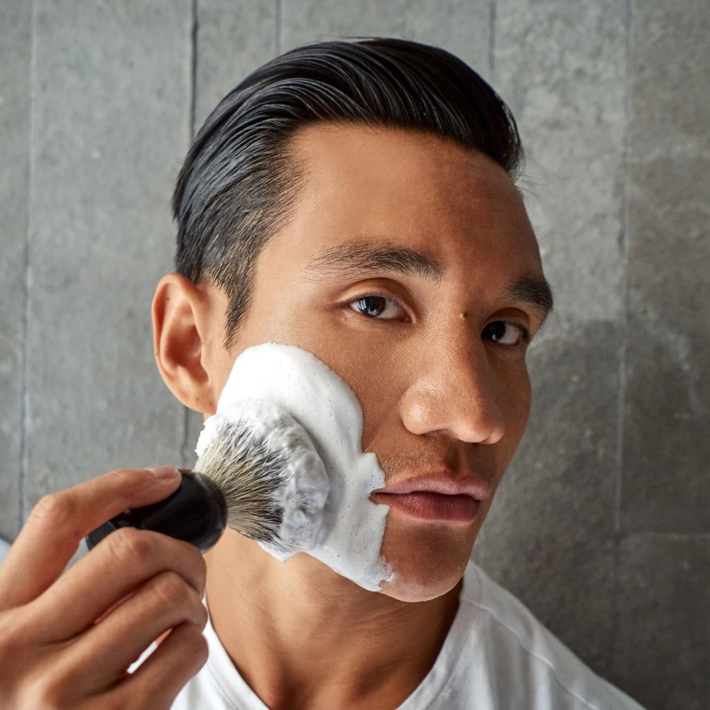 Mann rasiert sich mit Rasierpinsel und Schaum. Schwarzer Griff, Borsten auf der Wange. Grauer Hintergrund.