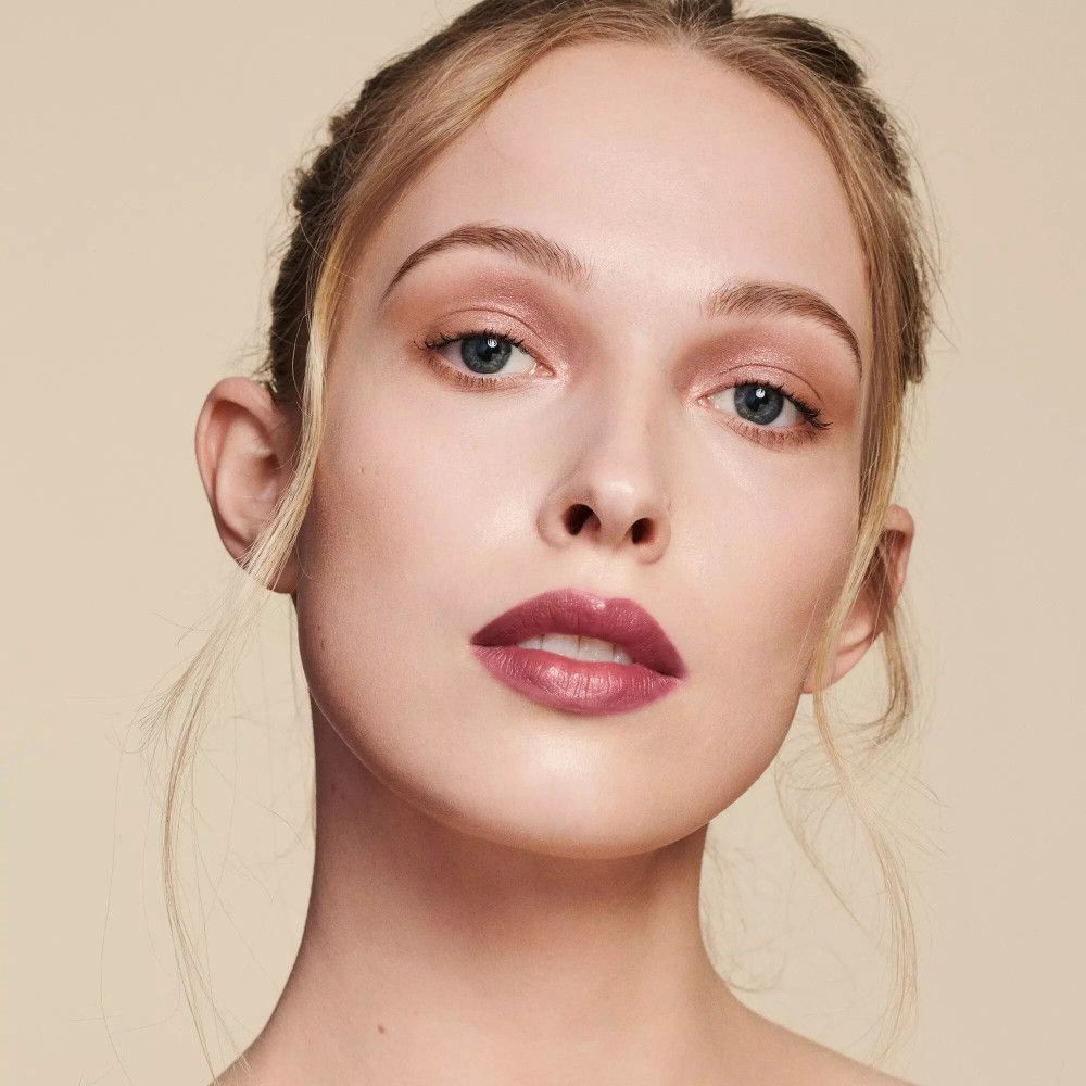 Nahaufnahme einer Frau mit hellem Teint und blonden Haaren. Sie trägt Make-up, einschließlich Lippenstift. Der Hintergrund ist hellbeige.