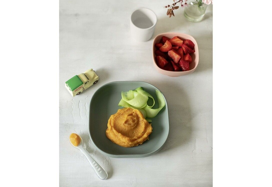 Grüner Teller mit Gemüse und Brei. Rosa Schüssel mit Erdbeeren. Weißer Becher. Spielzeugauto.