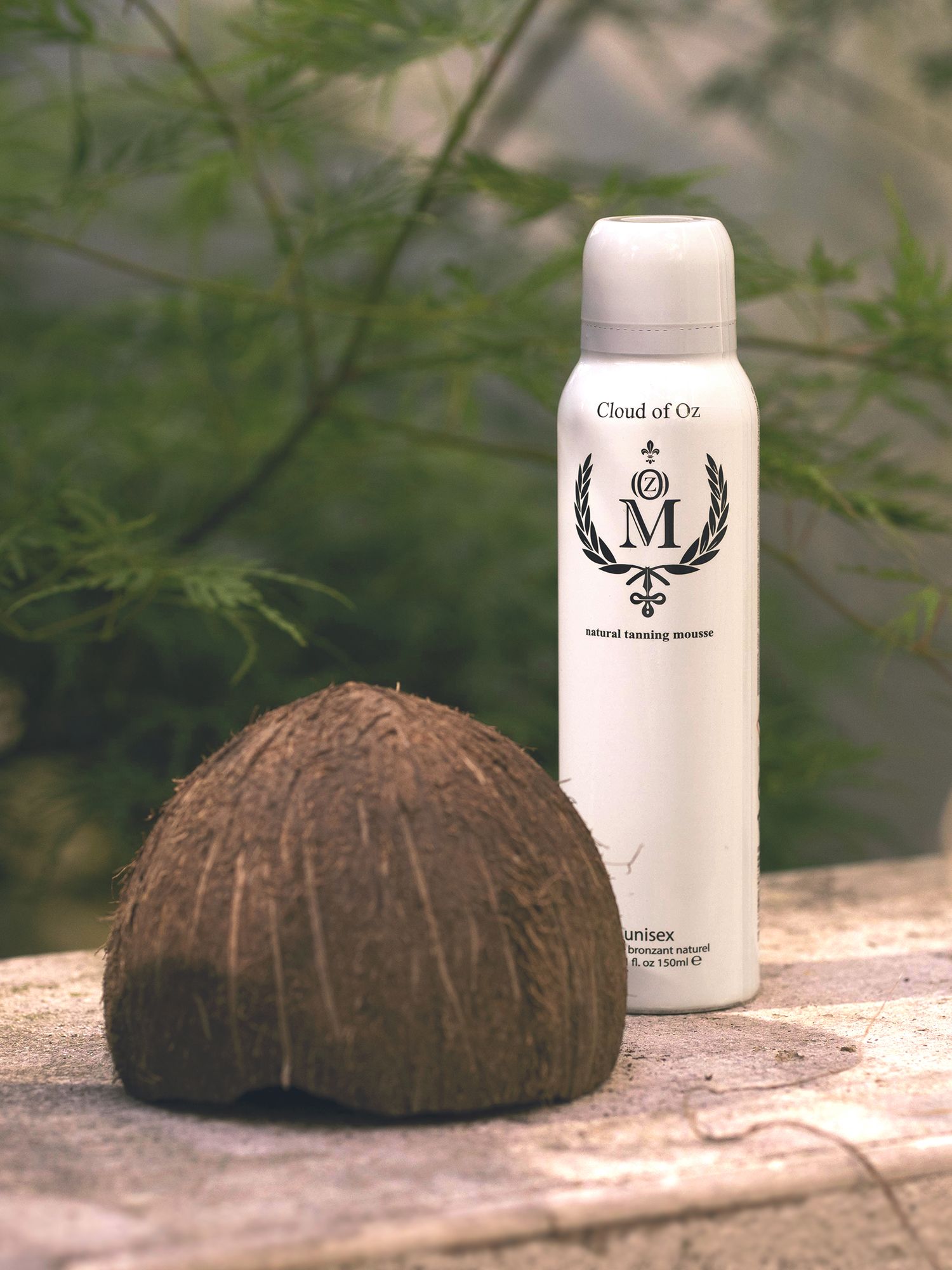 Weiße Sprühflasche mit schwarzem Logo und Schriftzug neben einer Kokosnussschale. Aufschrift: Cloud of Oz, Natural Tanning Mousse.