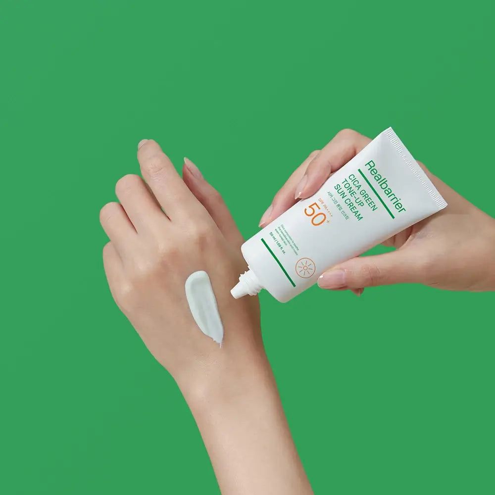 Hände halten Tube Real Barrier Cica Green Tone-Up Sun Cream. Creme wird auf die Hand aufgetragen.