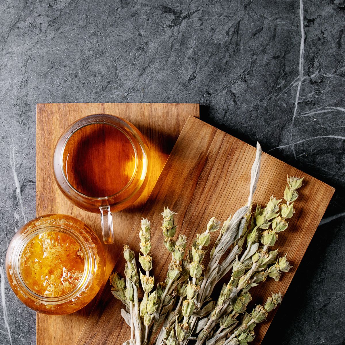 Glasbecher Tee, Honigglas, Bergtee-Bündel auf Holzbrett. Auf dunklem Steinuntergrund.