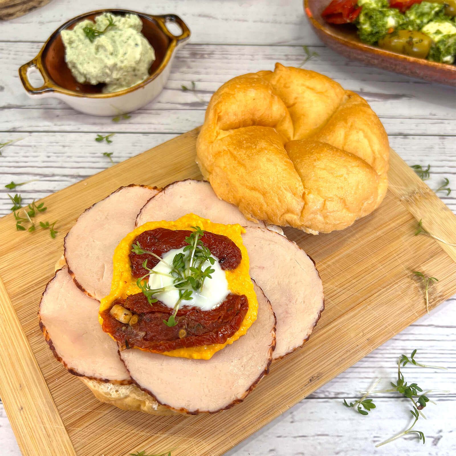 Kaiserbrötchen mit Belag auf einem Holzbrett. Belegt mit Fleisch, Tomaten, Kräutern und einem Dip. Daneben eine Schale mit Dip.