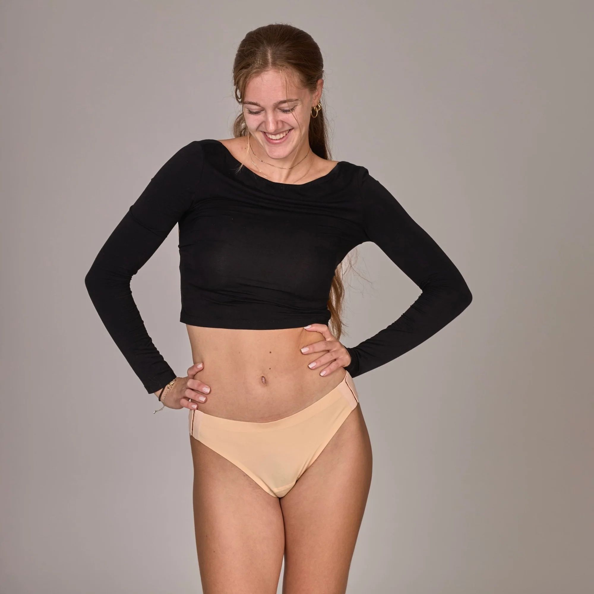 Frau in schwarzem Oberteil und haut farbenem Slip. Sie lächelt und steht mit den Händen auf den Hüften.
