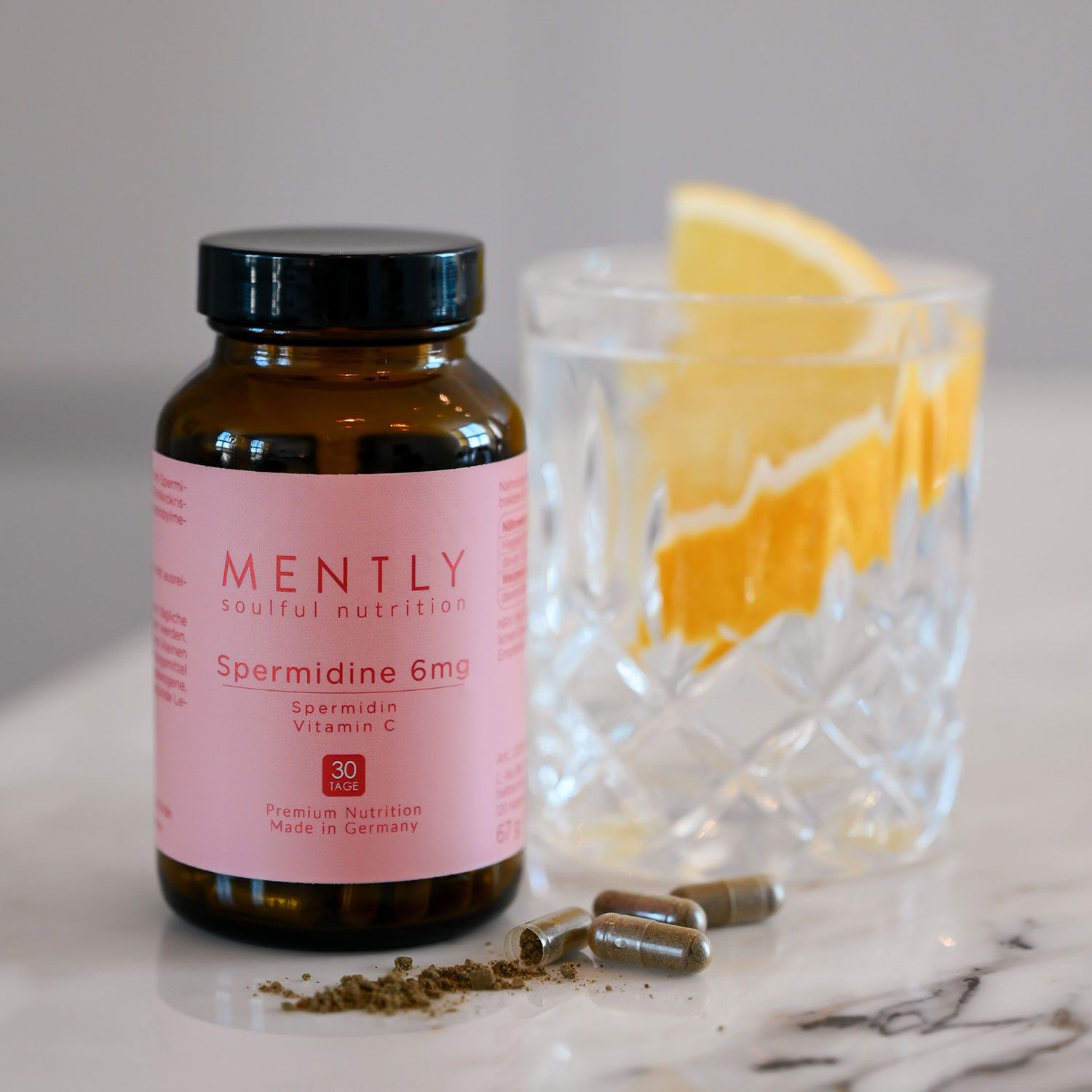 Braune Glasflasche mit Kapseln, daneben Glas mit Wasser und Zitrone. Aufschrift "MENTLY Spermidine 6mg".