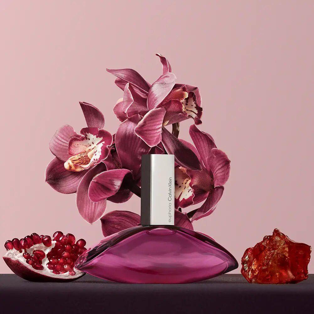 Euphoria-Flakon mit silbernem Verschluss, umgeben von Orchideen, Granatapfel und einem bernsteinfarbenen Objekt. Rosa Hintergrund.