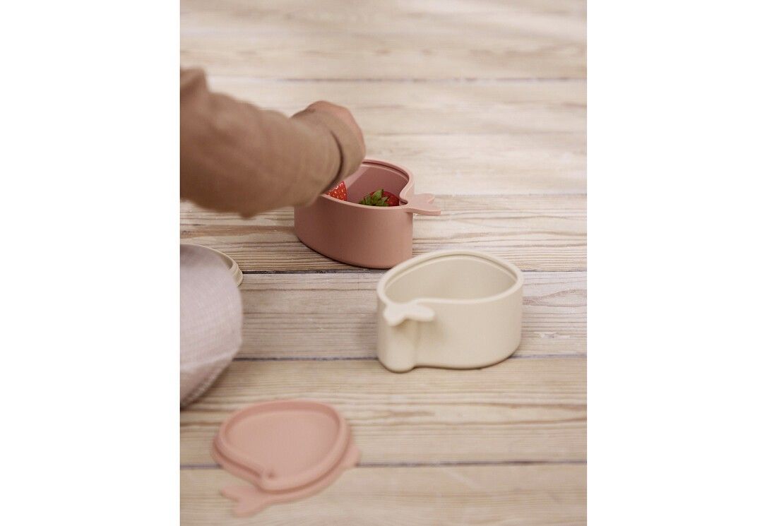 Zwei walförmige Brotdosen auf Holzuntergrund. Eine Dose geöffnet, eine Hand greift hinein. Rosa und Beige.