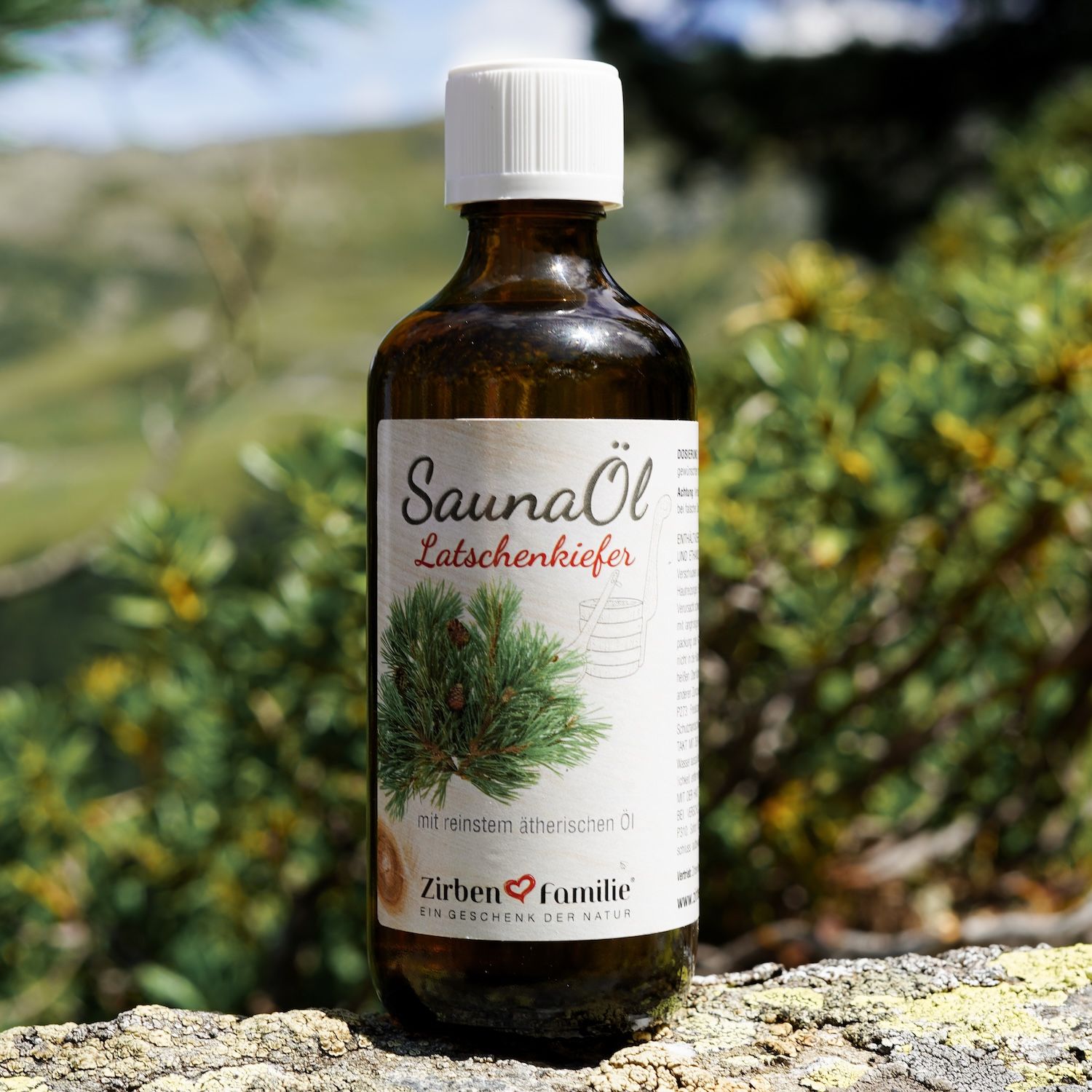 Braune Glasflasche mit weißem Deckel. Etikett mit SaunaÖl Latschenkiefer und ZirbenFamilie-Logo. Im Hintergrund: grüne Pflanzen und Felsen.