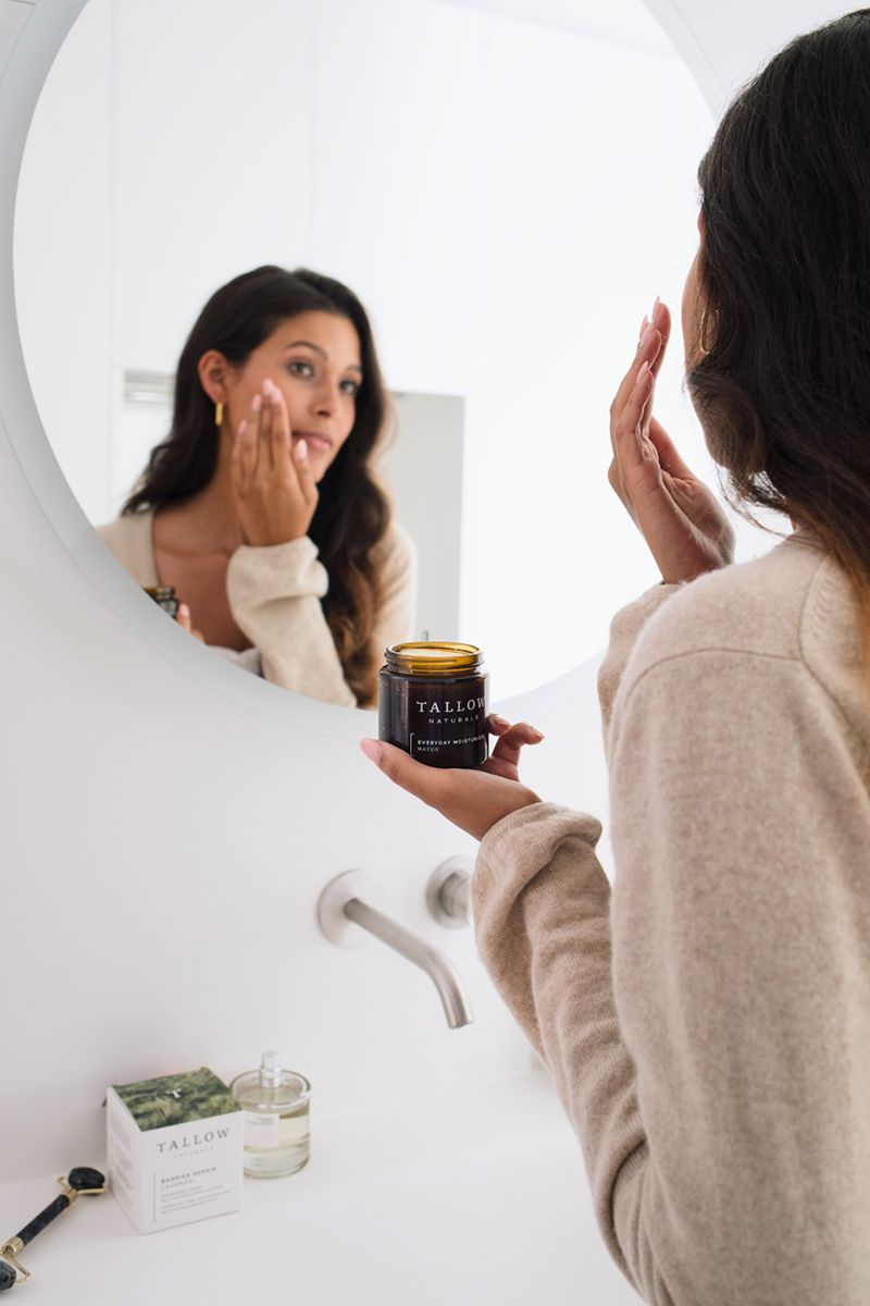 Frau betrachtet sich im Spiegel. Hält einen Tiegel mit Aufschrift Tallow Naturals. Weitere Produkte auf der Ablage.
