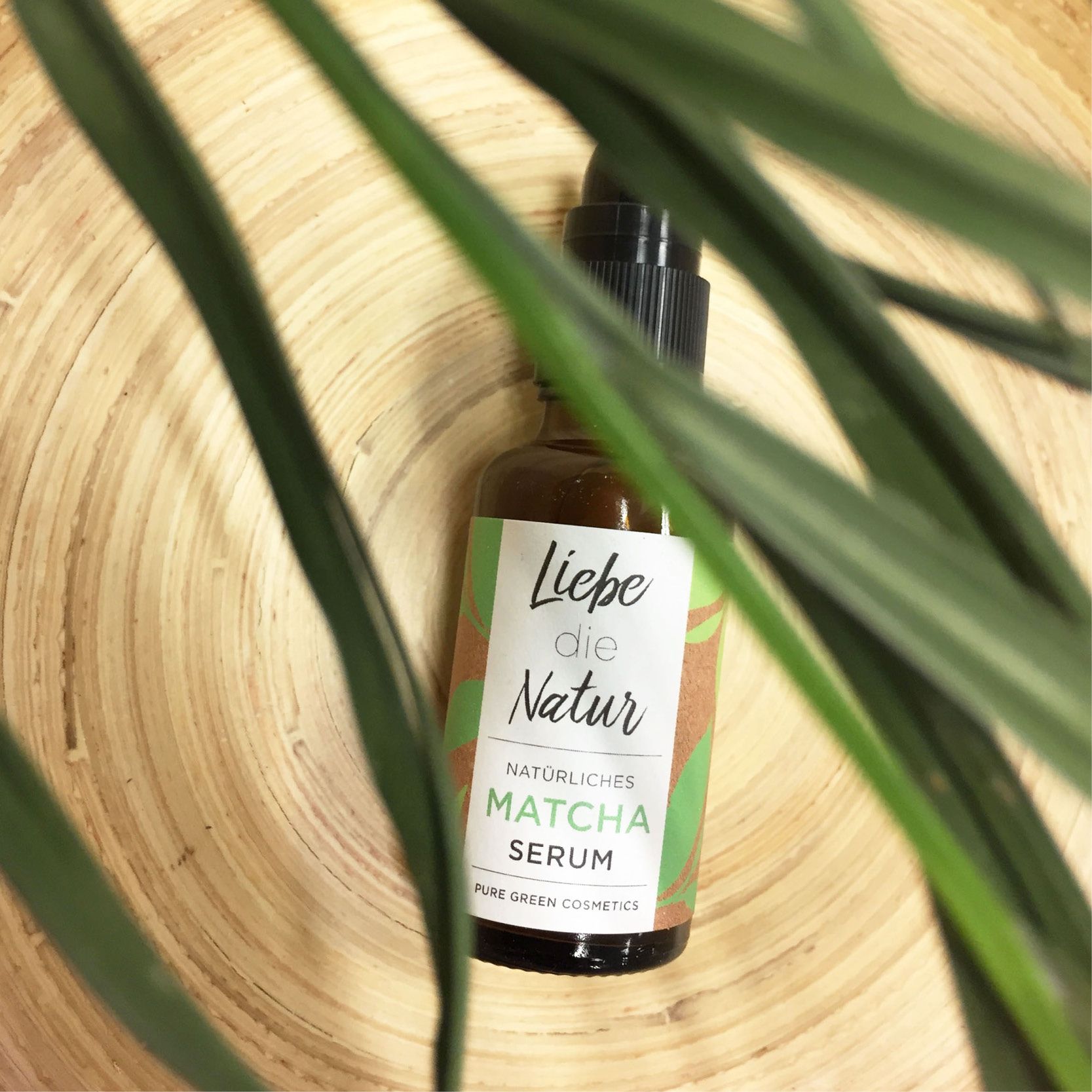 Braune Glasflasche mit schwarzem Sprühkopf. Etikett mit "Liebe die Natur" und "Matcha Serum". Grüne Blätter im Vordergrund.