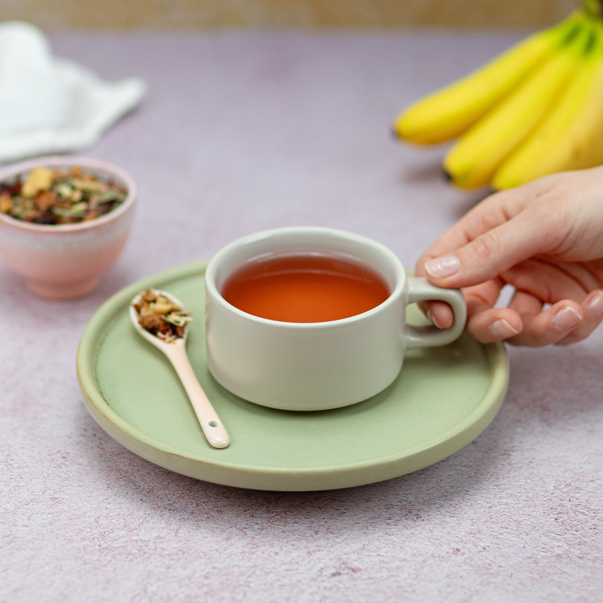 Tasse Tee auf Untertasse. Löffel mit losem Tee, Schale mit Tee. Hand hält Tasse. Hintergrund: Teller, Bananen.