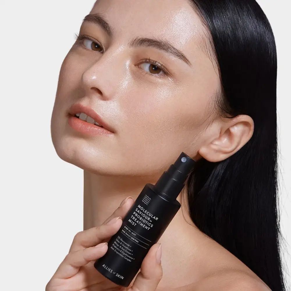 Eine Frau hält eine schwarze Sprühflasche. Auf der Flasche steht: MOLECULAR SAVIOUR PROBIOTICS TREATMENT MIST. Sie sprüht das Produkt auf ihr Gesicht.