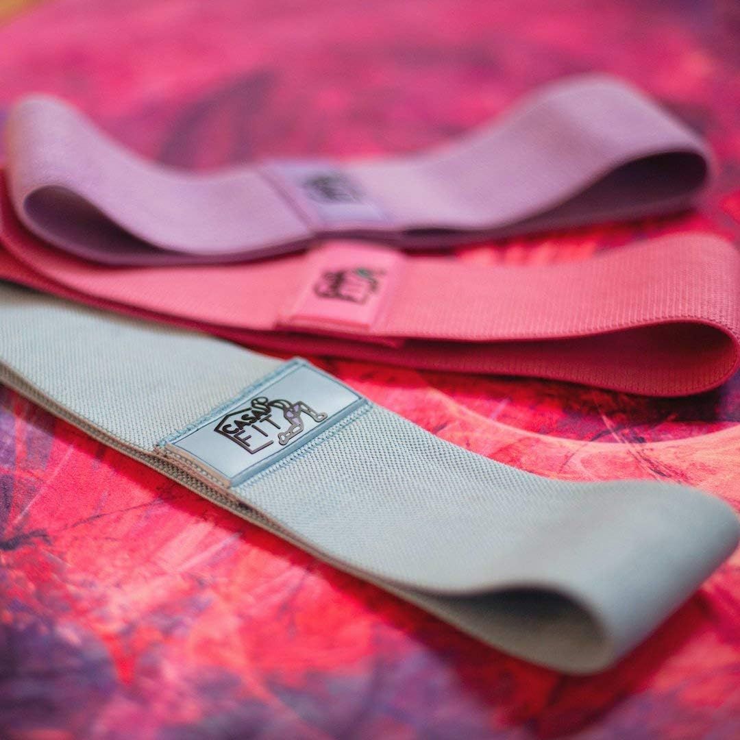 Drei Hüftbänder in Pastellfarben: Lila, Rosa und Hellblau. Jedes Band hat ein rechteckiges Label mit Logo.