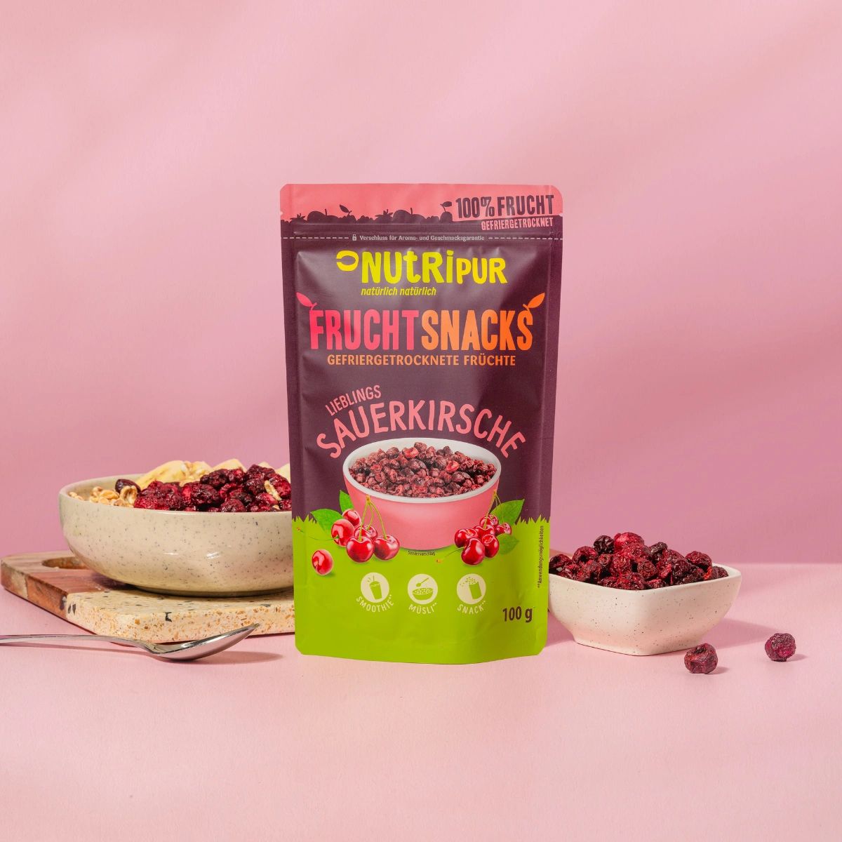 Schale mit Müsli, Bananenscheiben und gefriergetrockneten Sauerkirschen. Daneben Beutel mit Sauerkirschen. Rosa Hintergrund.