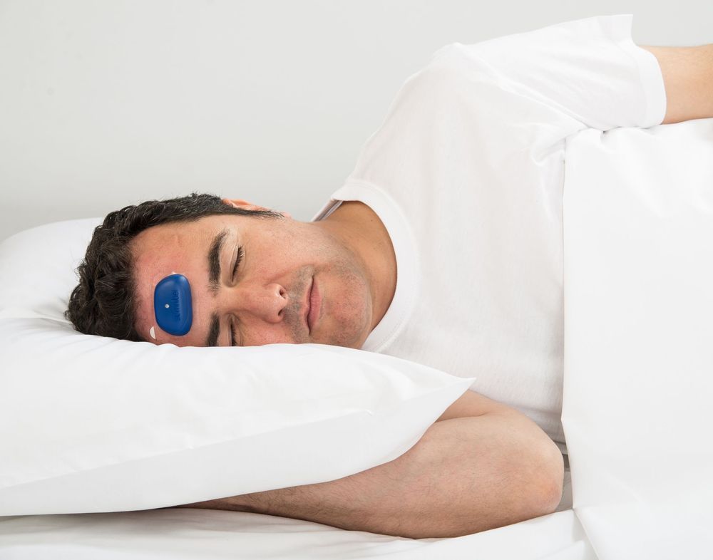 Mann im Bett mit blauem Gerät auf der Stirn. Liegt auf einem Kissen.