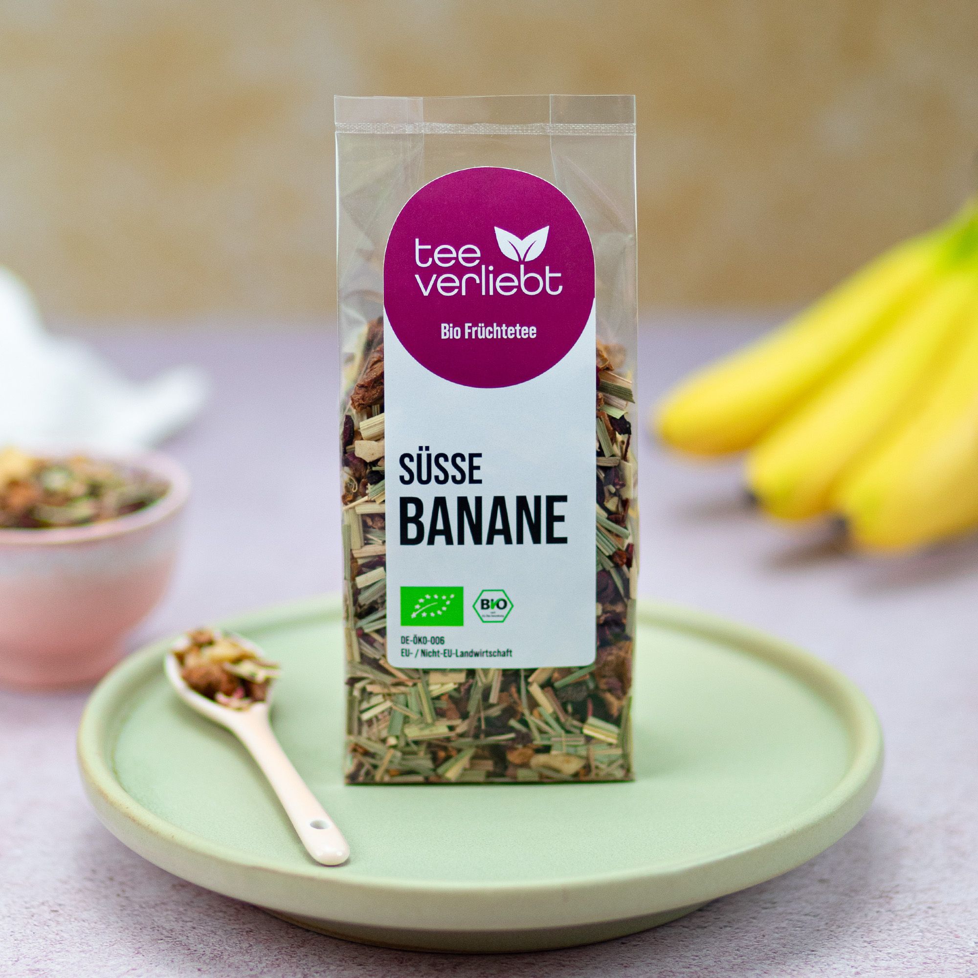 Klarsichtbeutel mit losem Früchtetee auf Teller. Logo, Text 'Süße Banane' und Bio-Siegel. Bananen im Hintergrund, Löffel mit Tee.