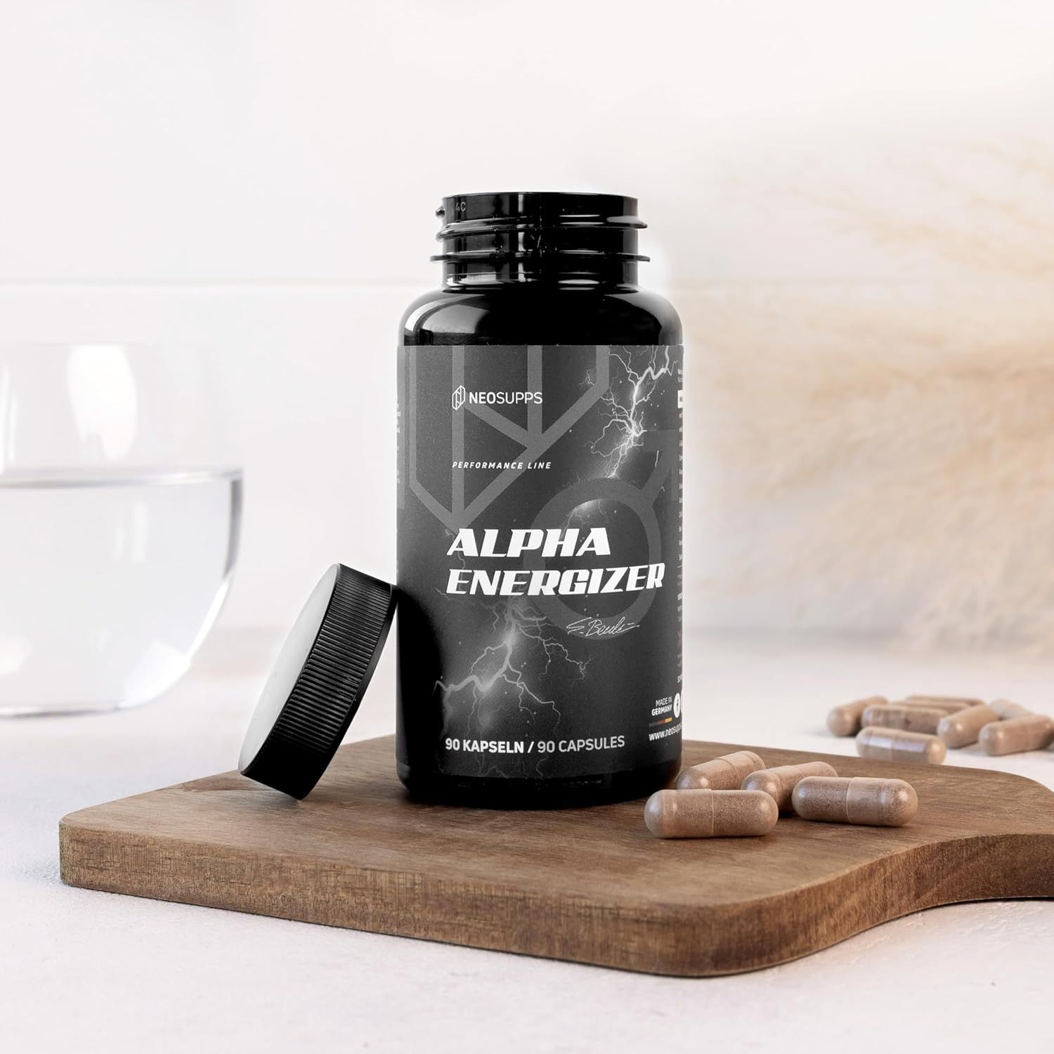 Schwarze Flasche "Alpha Energizer" mit geöffnetem Deckel und Kapseln auf Holzbrett. Wasserglas daneben.