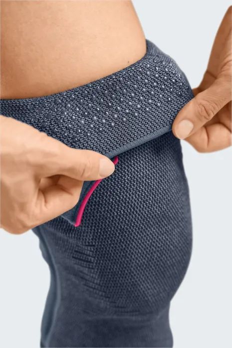 Nahaufnahme einer Kniebandage. Hände ziehen einen Teil der Bandage hoch. Graues Material mit Noppen.