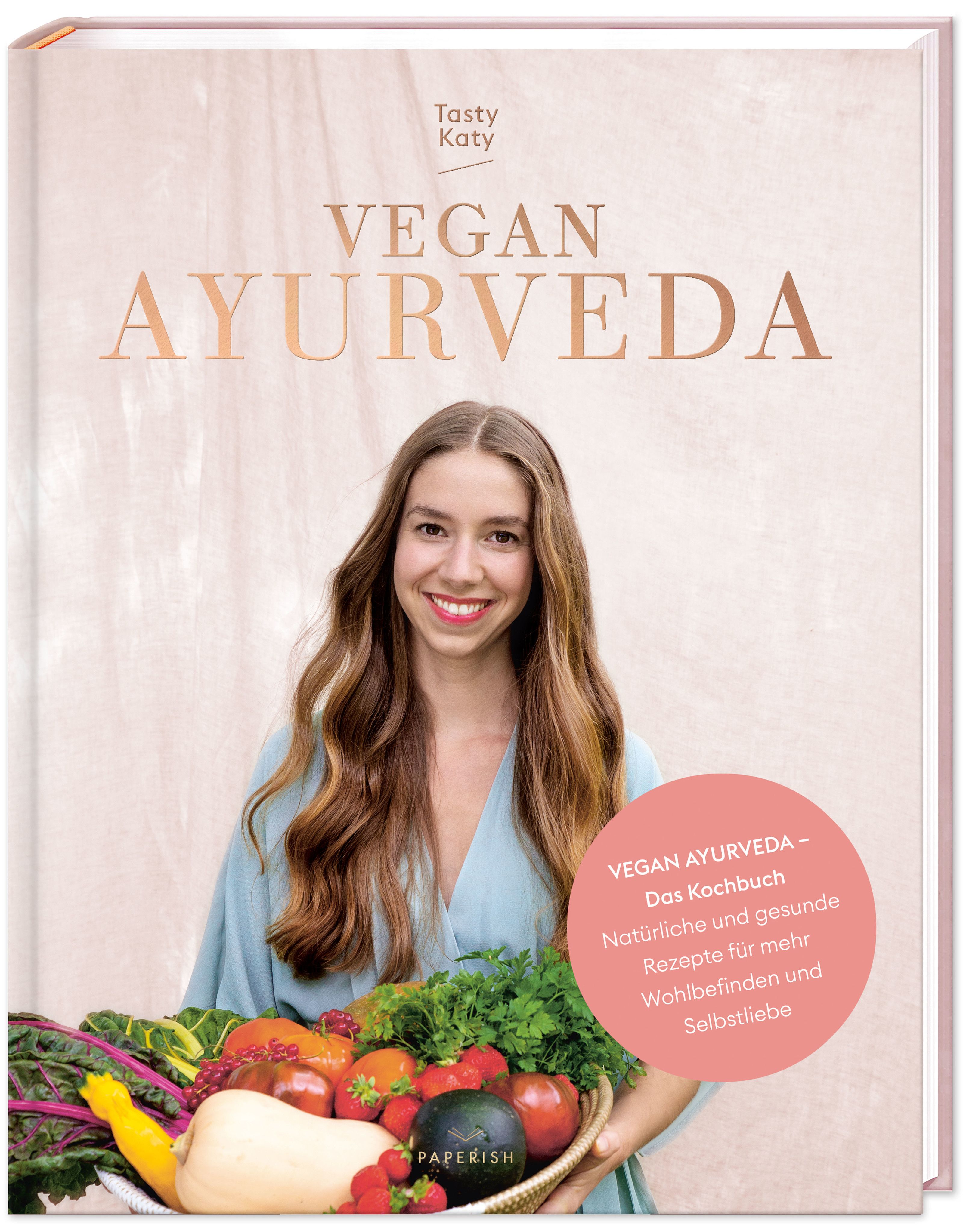Buch mit dem Titel VEGAN AYURVEDA. Auf dem Cover ist eine Frau mit Gemüsekorb abgebildet. Rosa Kreis mit Text: Vegan Ayurveda - Das Kochbuch.