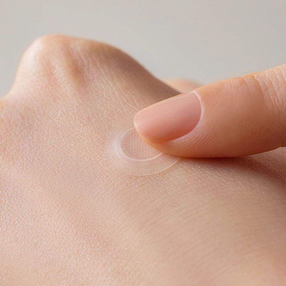 Ein Finger platziert einen transparenten, runden Patch auf der Haut. Nahaufnahme einer Hand.