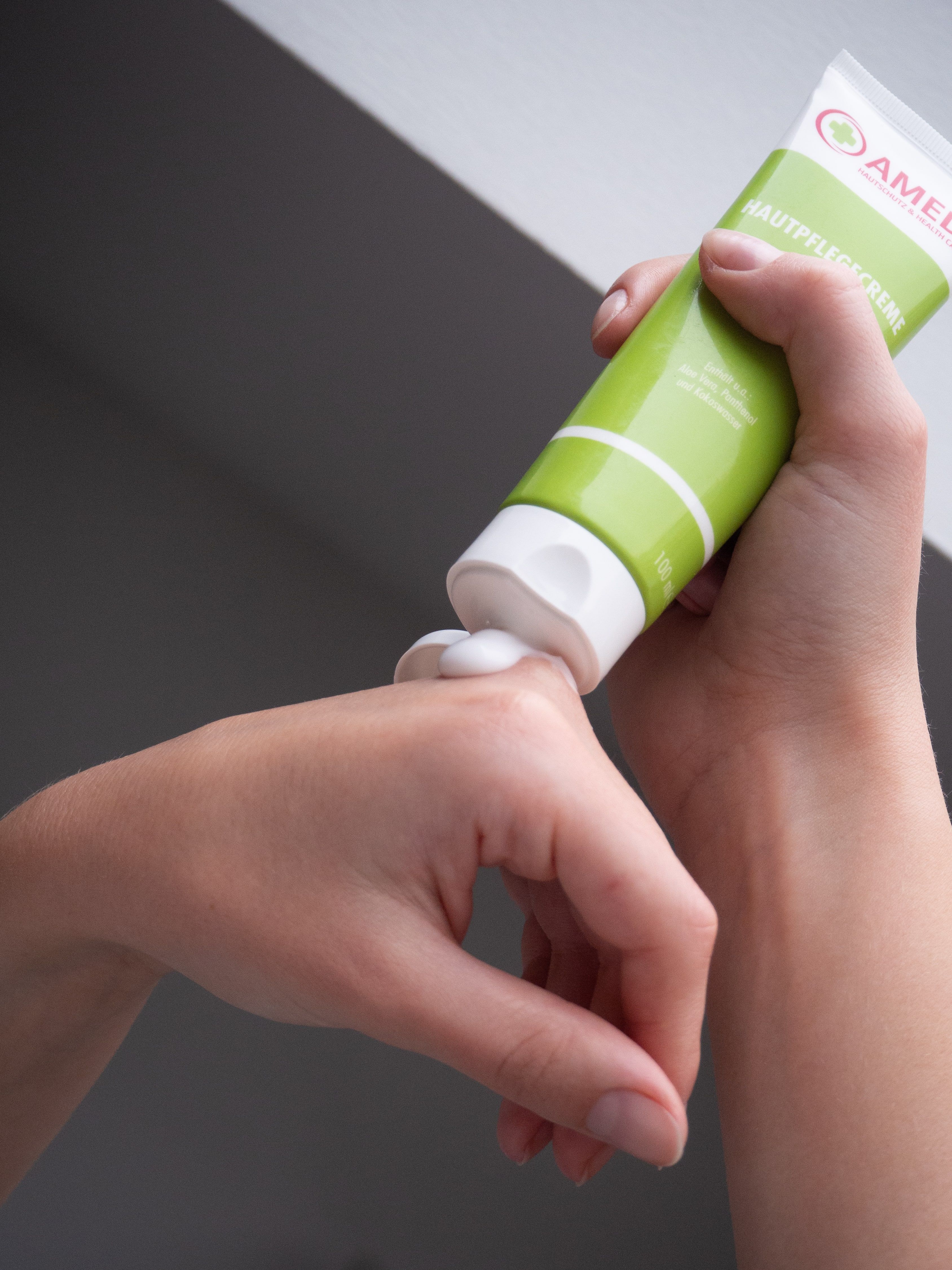 Hand drückt Creme aus grüner Tube. Aufschrift: AMEDI, Hautpflegecreme. Logo mit rotem Kreuz. Text in Deutsch.